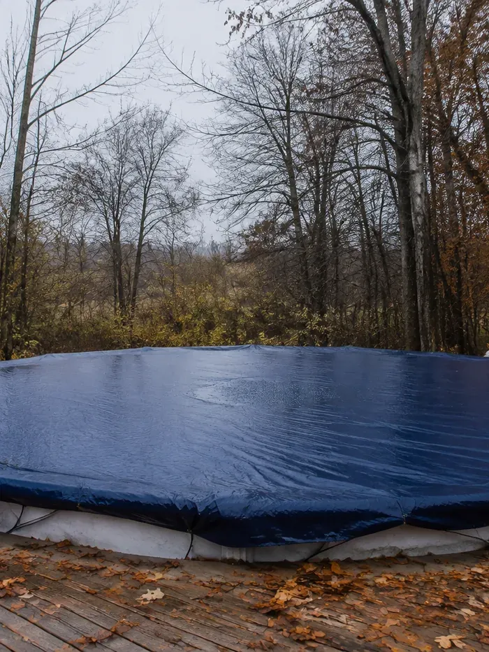 Bâche d'hivernage bleu foncé tendue sur une piscine ronde, terrasse en bois jonchée de feuilles mortes