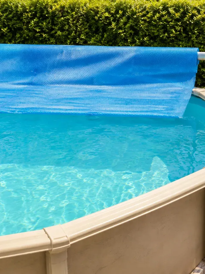 Bâche à bulles bleue déroulée sur une piscine hors sol à l'eau turquoise, devant une haie
